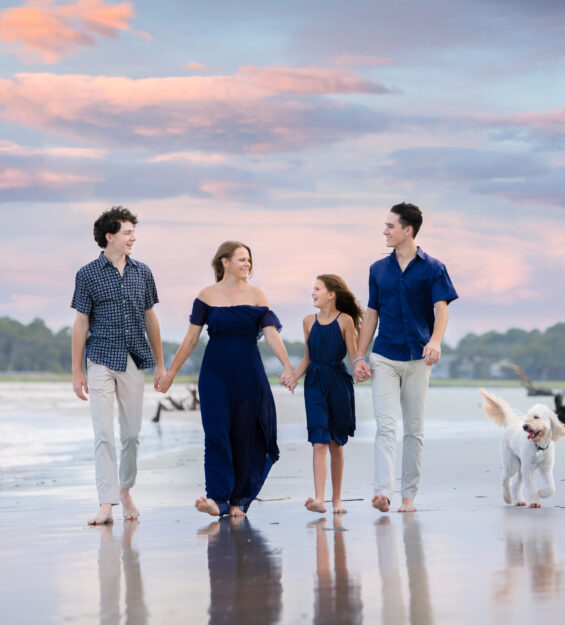 Family beach portrait session during sunset with pastel skies. This photo captures a family enjoying a barefoot walk with their dog along the shore. Ideal for those searching for ‘beach family photoshoot ideas’ or ‘sunset family portraits