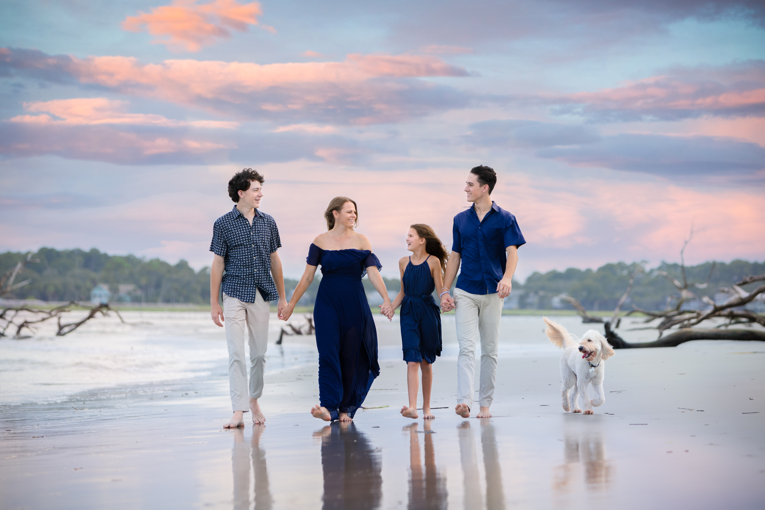 Family beach portrait session during sunset with pastel skies. This photo captures a family enjoying a barefoot walk with their dog along the shore. Ideal for those searching for ‘beach family photoshoot ideas’ or ‘sunset family portraits